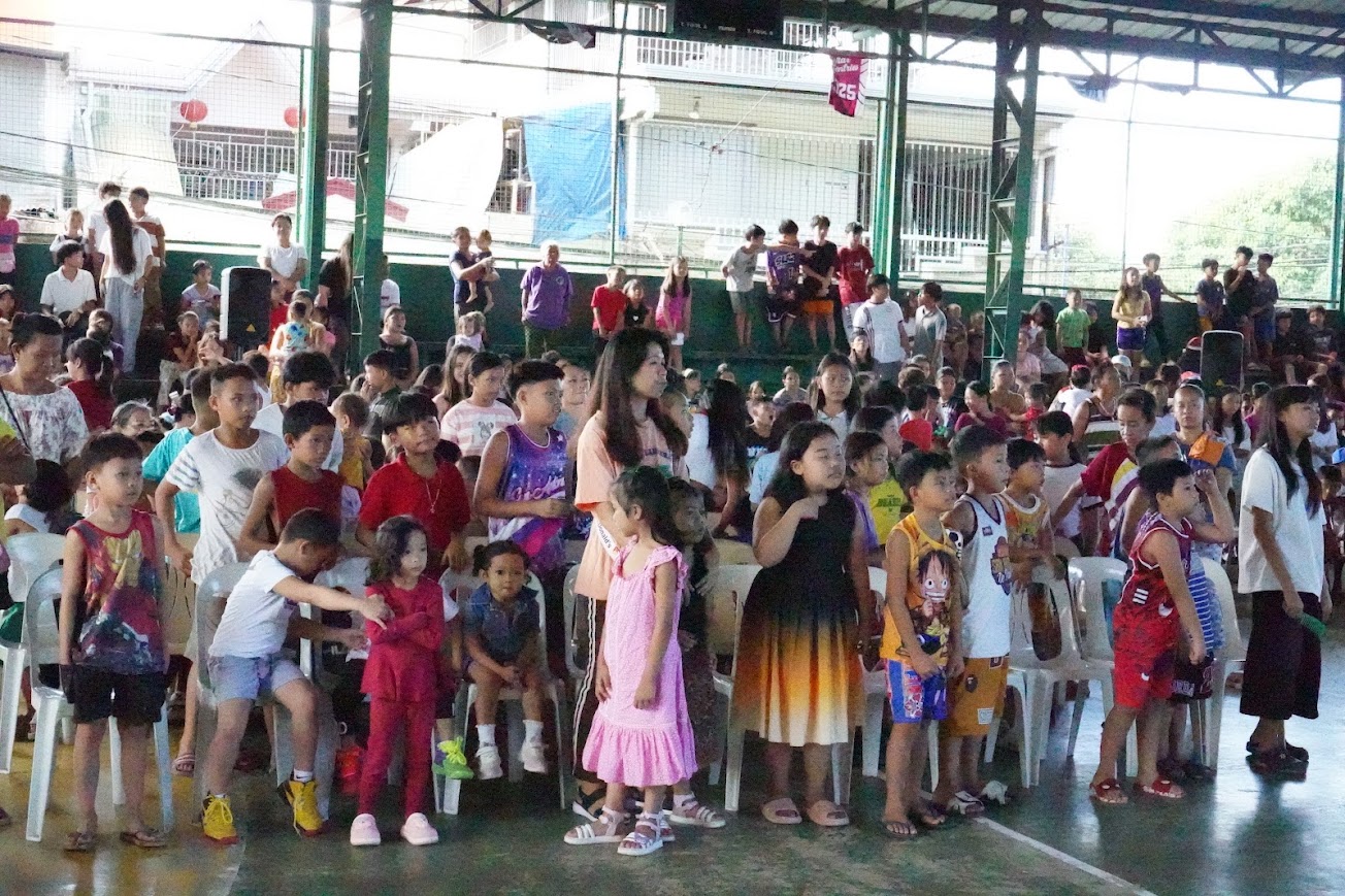 Children stand together in the front rows, morning sun through the roof.