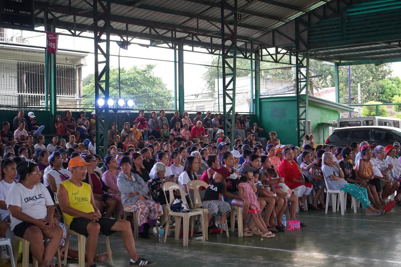 A team member speaks to the seated crowd, jeepneys parked at the far end.