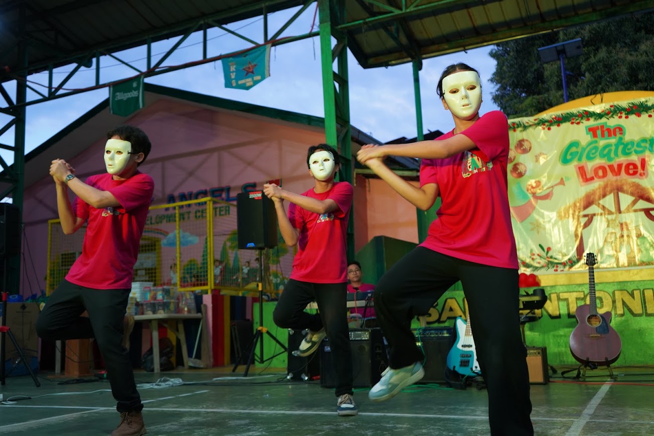 The youth dance team performs in masks, "The Greatest Love!" banner behind them.