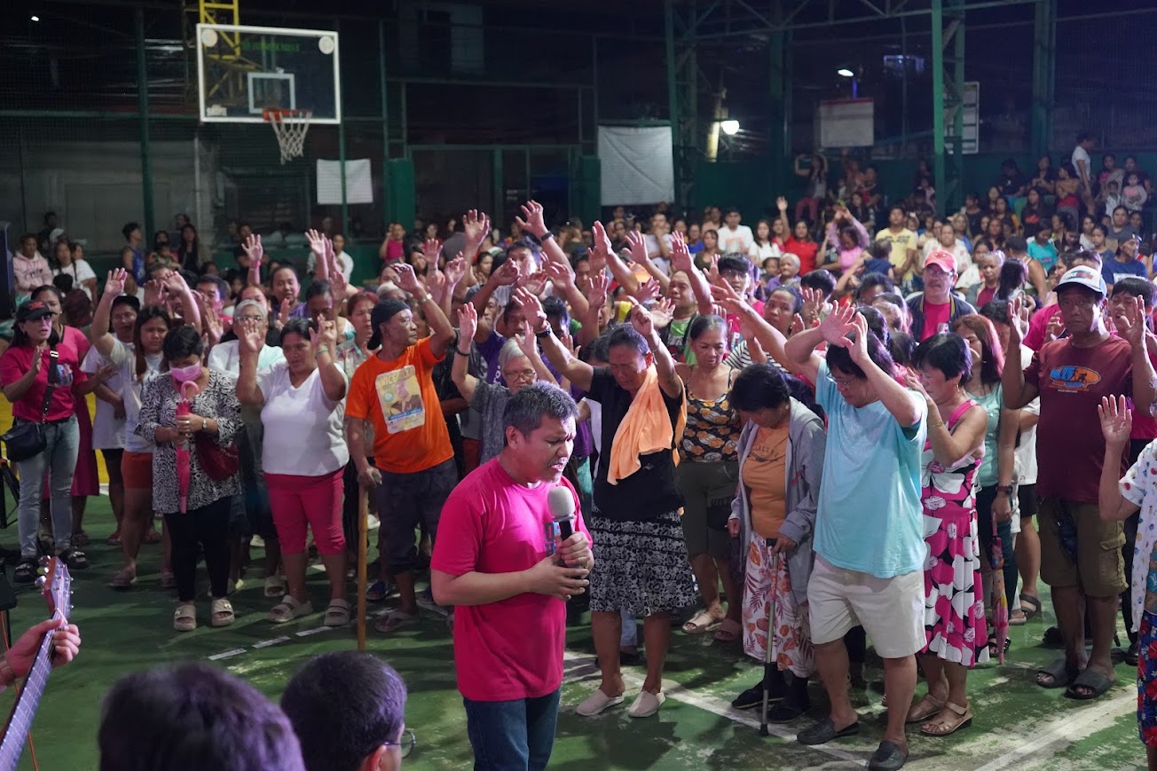 A team member at the mic, head bowed, the crowd with hands lifted behind him.