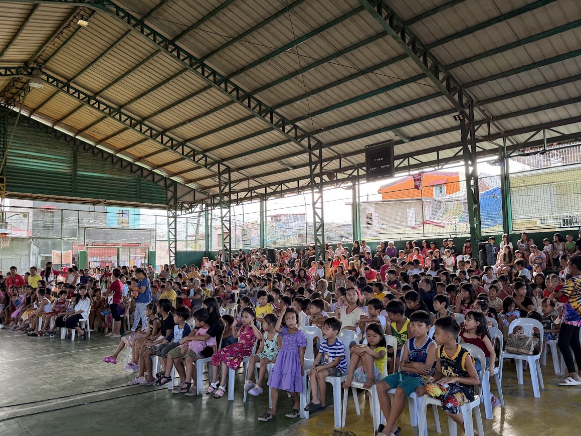 The court fills — rows of children seated, families in the stands behind.