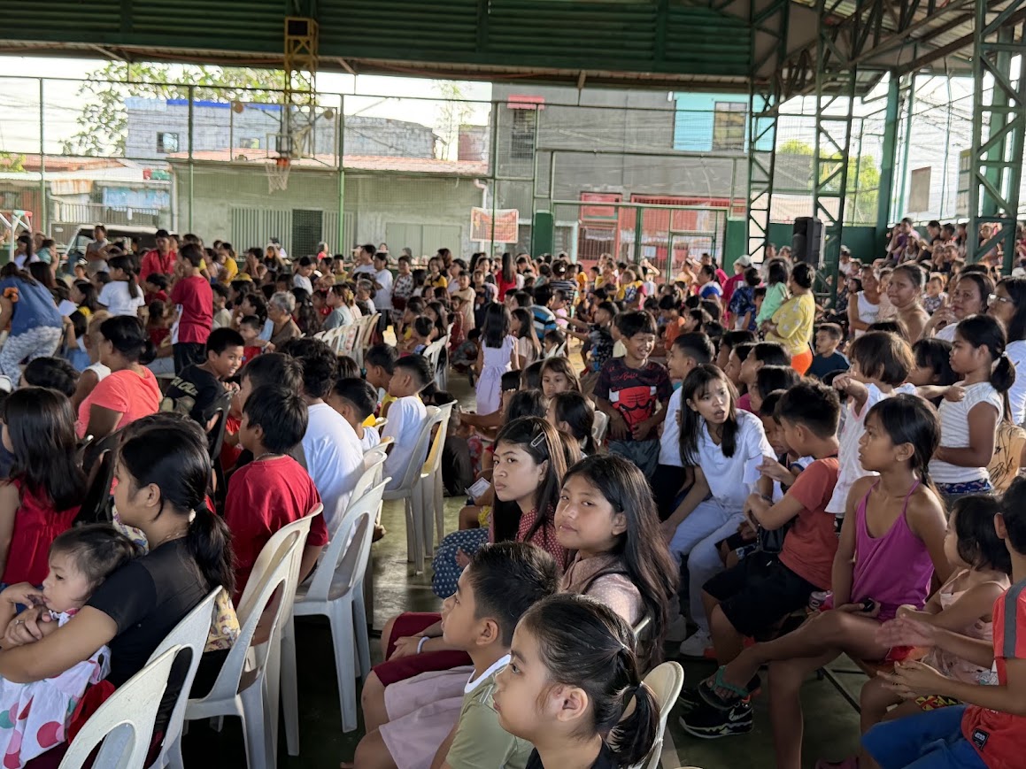 Children seated in the front rows, morning sun on the far side of the court.