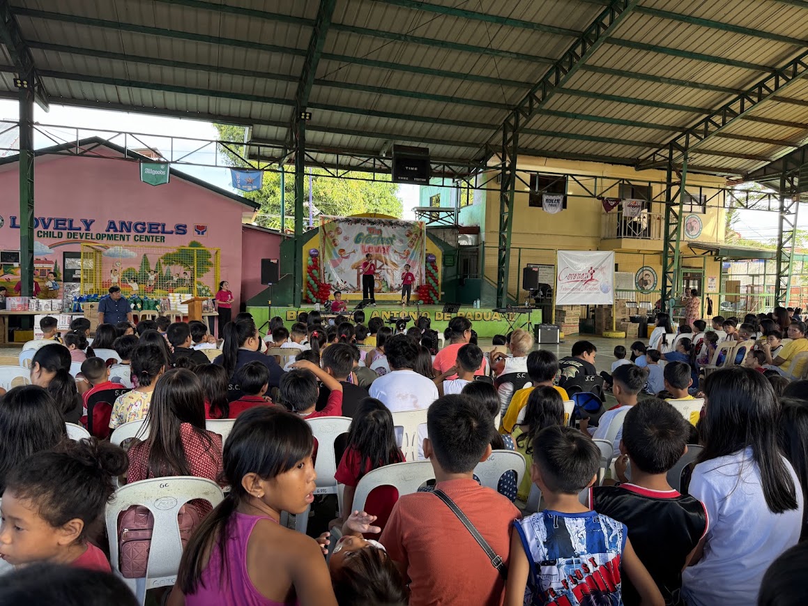 From the back — children facing "The Greatest Love!" stage, the Covenant banner at right.
