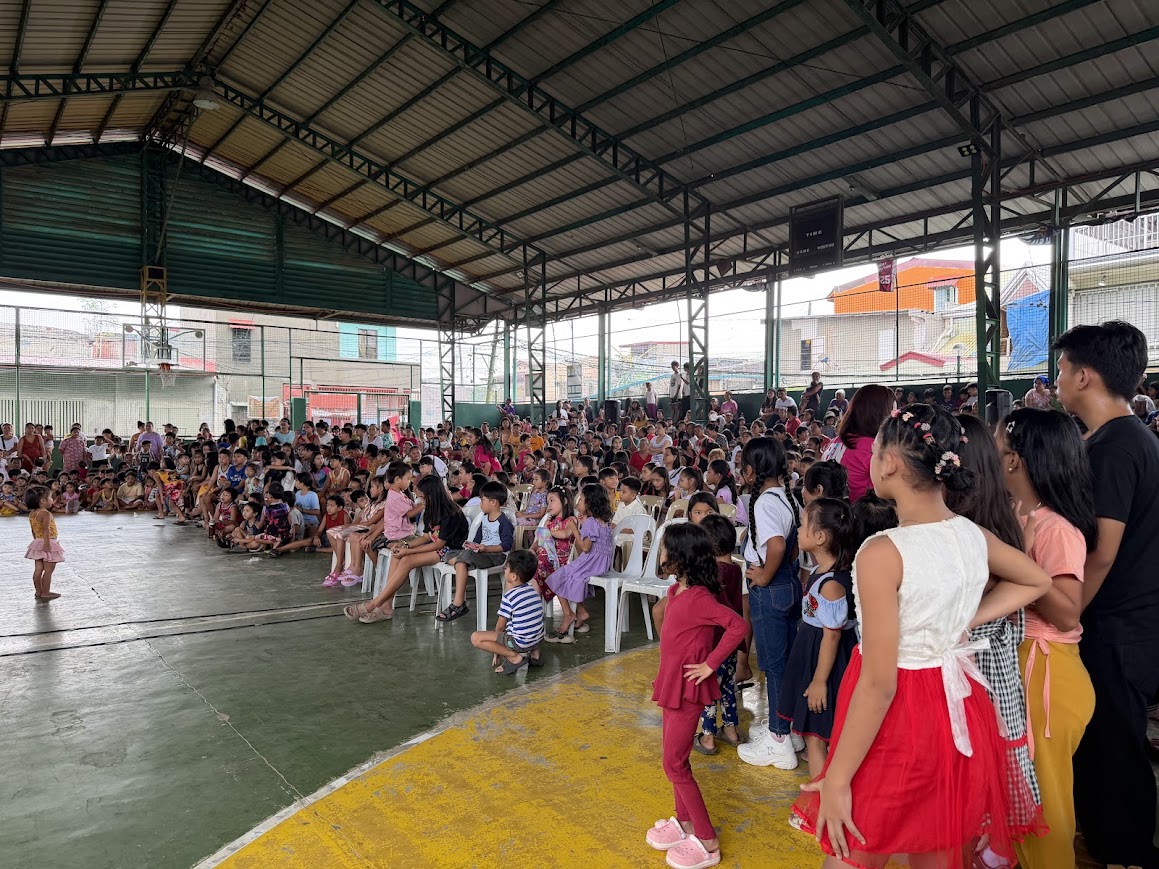 Children line the edge of the court as a small girl watches the performance from the open floor.