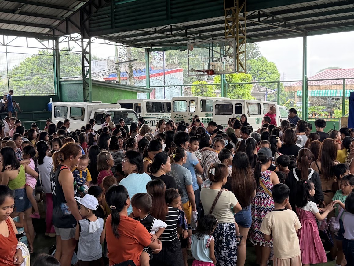 The crowd seen from behind the gift table, jeepneys parked at the far end.