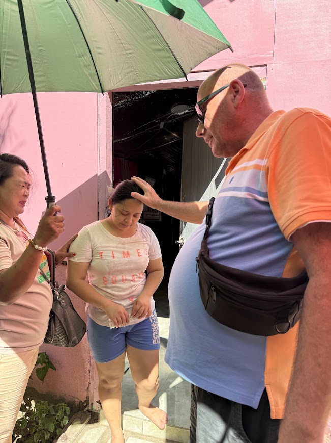 Prayer offered at a doorway during the morning outreach, shade held against the midday sun.