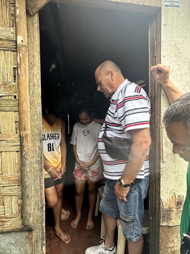 Morning door-to-door — a team member prays with a mother at her doorway, a hand on her forehead.