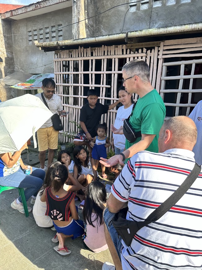 A team member speaks with the children seated in front of a bamboo-slat house.