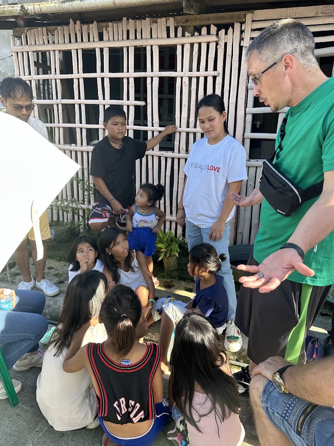 The team gathers with the family — children seated, adults standing at the house.