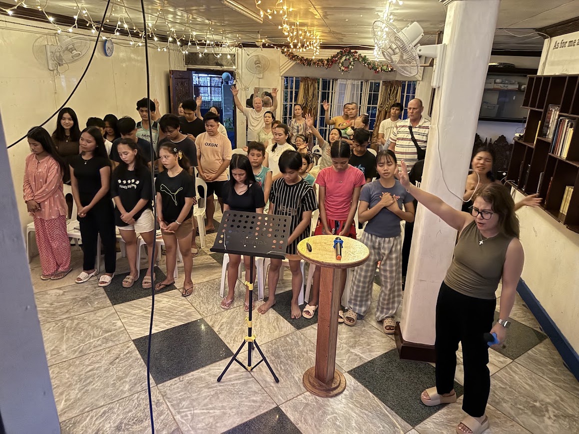 A team member leads prayer from the front as the congregation stands, heads bowed.