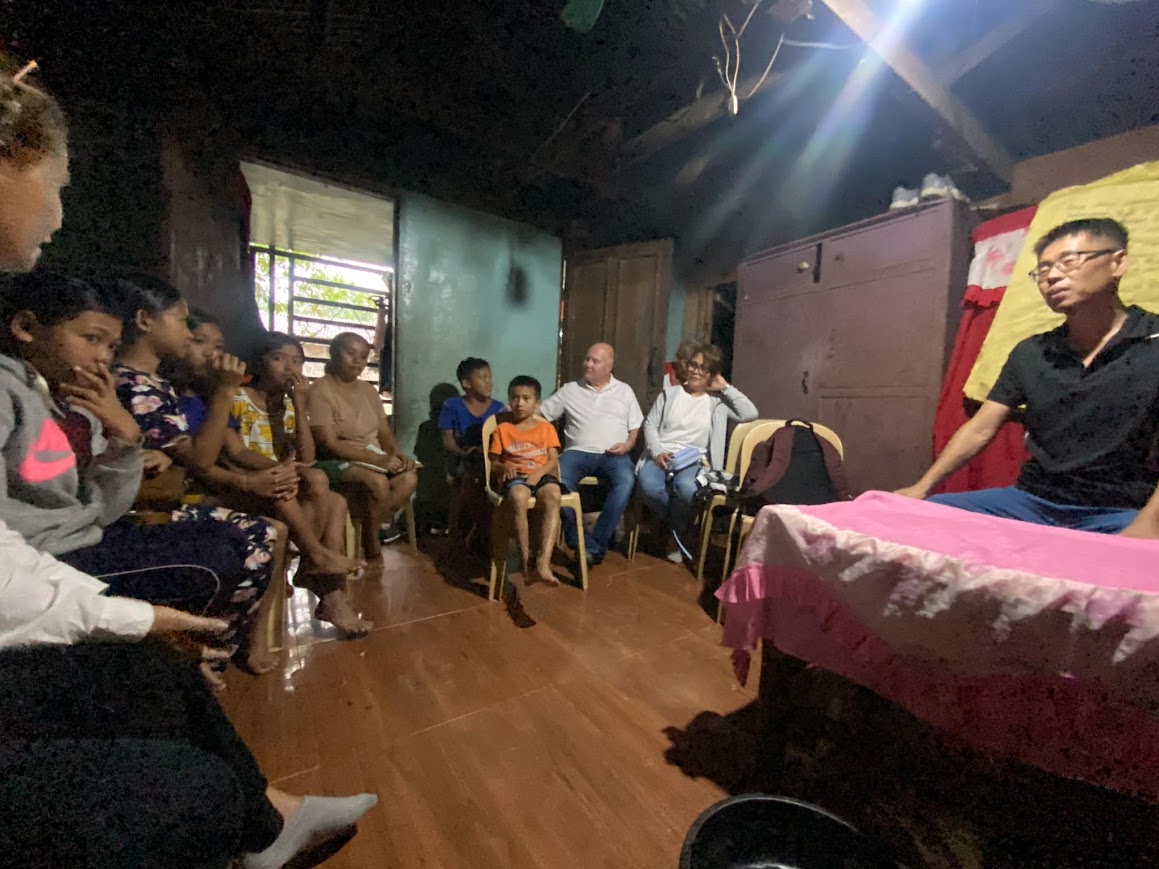 Inside a family's home — the team and family seated in a circle, a leader speaking from the bed.