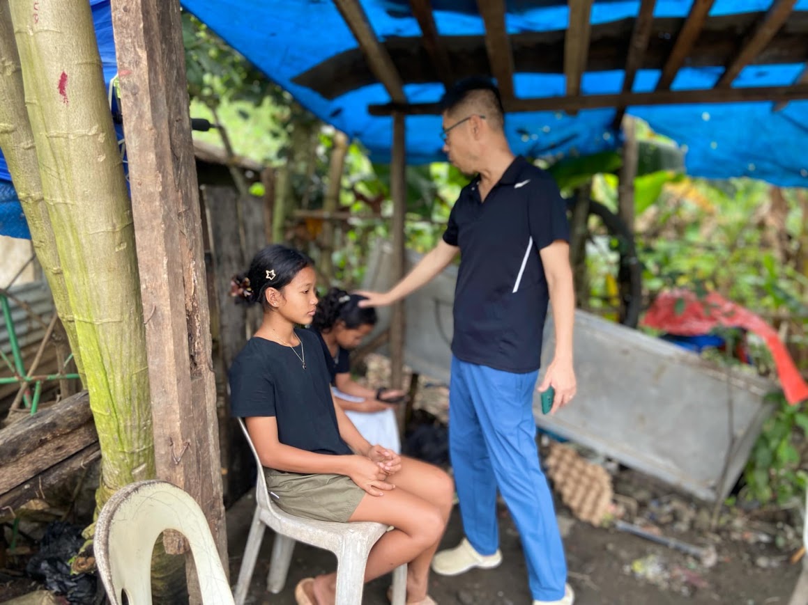 Under a tarp in the yard — a leader lays a hand on a seated girl's head.