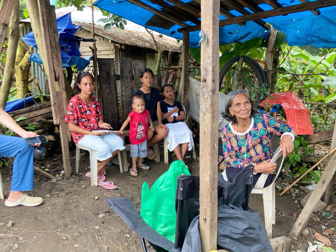 Under the tarp shelter — an elder with her granddaughters and a small boy, a team member seated beside them.