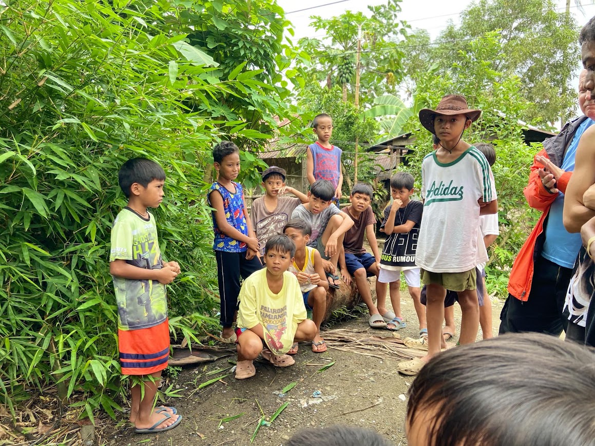 The boys gather at the edge of the bamboo, one in a bush hat, as the team arrives.