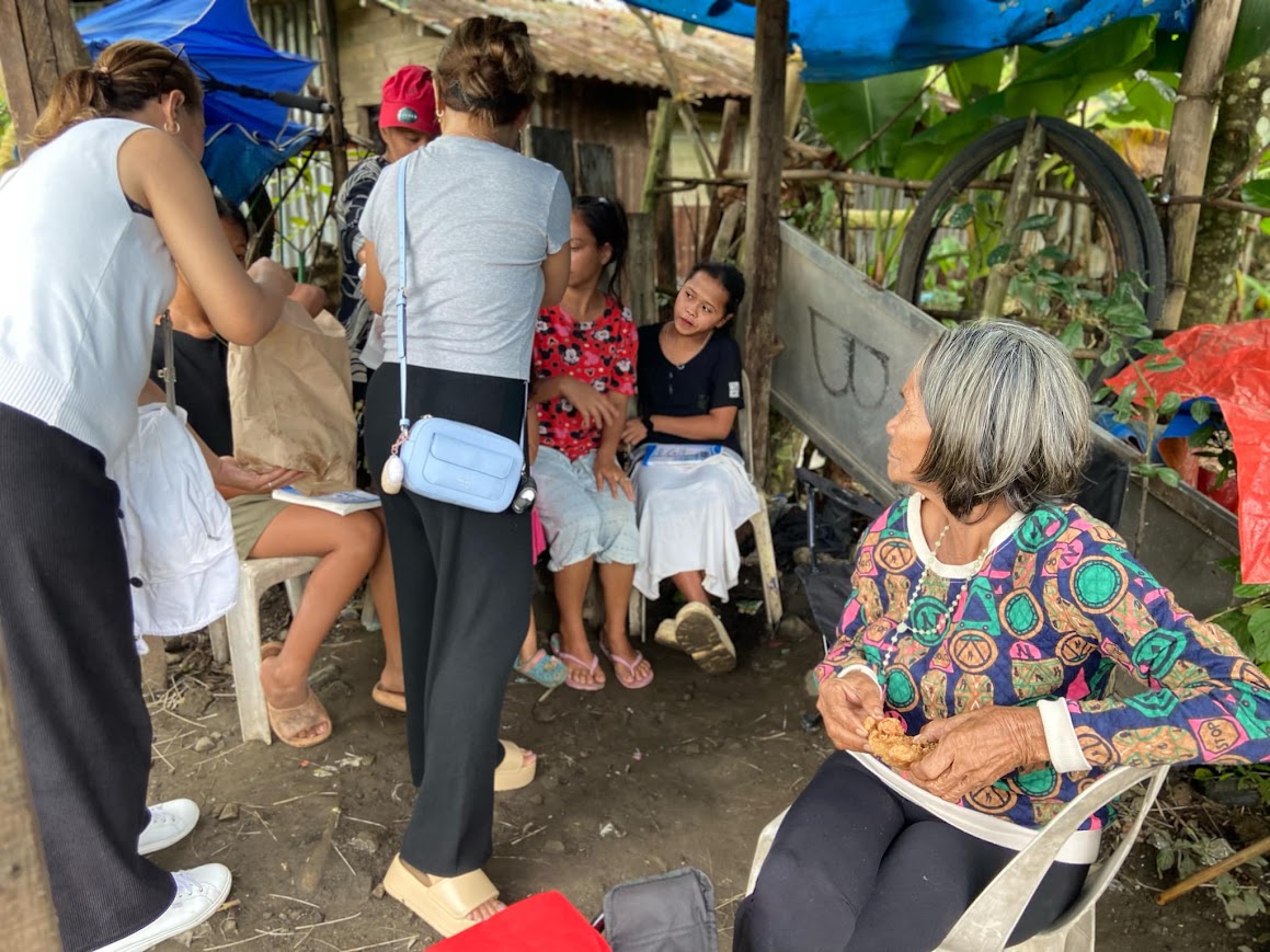 The team brings bread to the family under the tarp, the elder seated at the front.