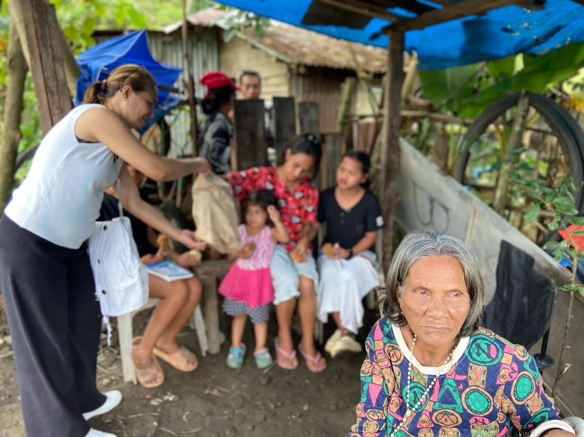 Bread handed to the family under the tarp, the elder seated in the foreground.