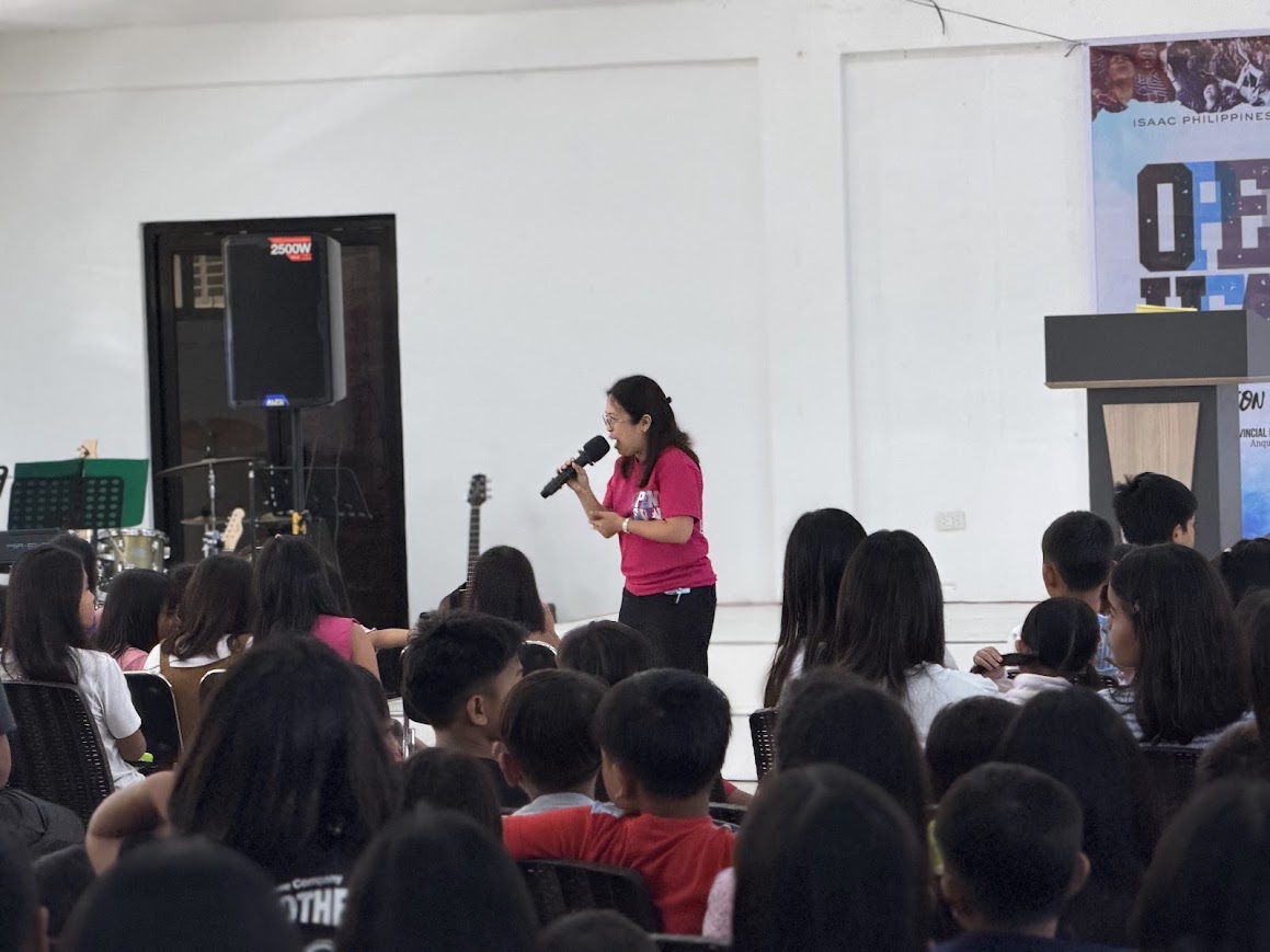 A team member speaks to the children from the front of the stage.