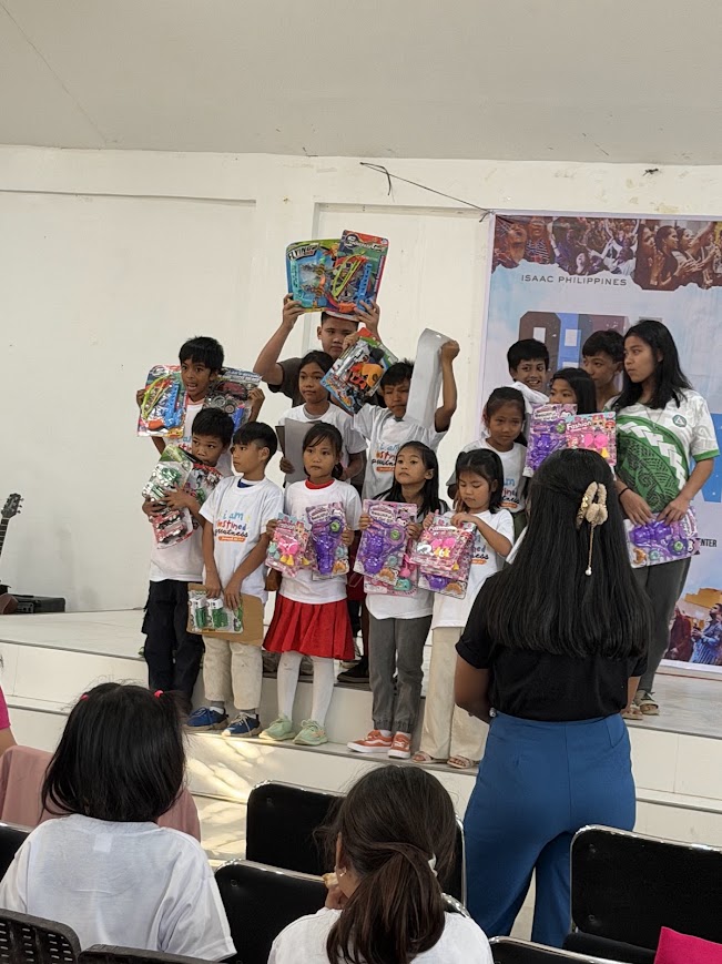 Children on stage with their gift packs, a volunteer holding one high.