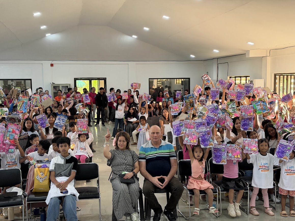 The full hall — every child holding up a gift pack, leaders seated at the center.