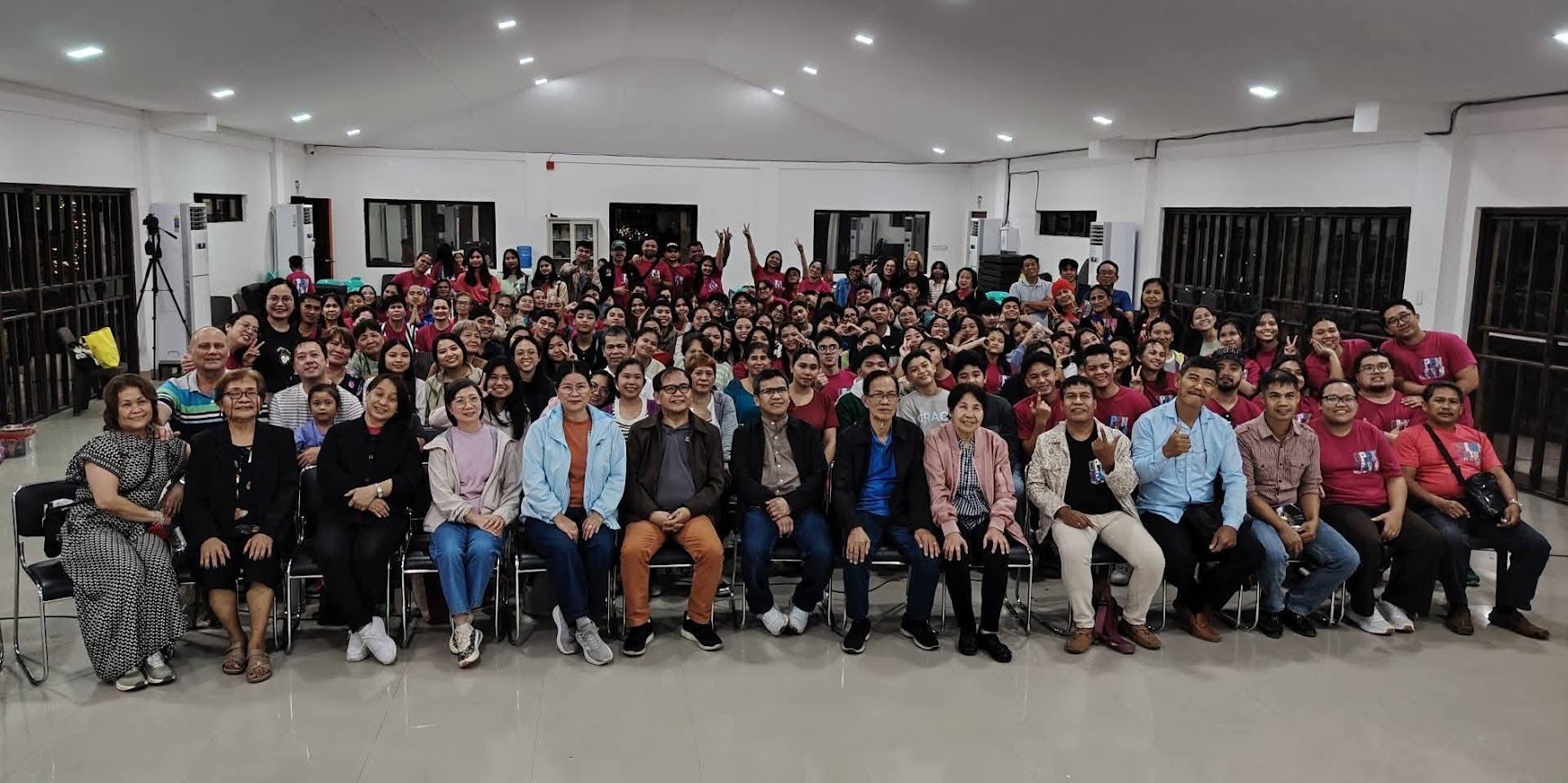 The full gathering — over a hundred people in the hall, leaders seated in front.