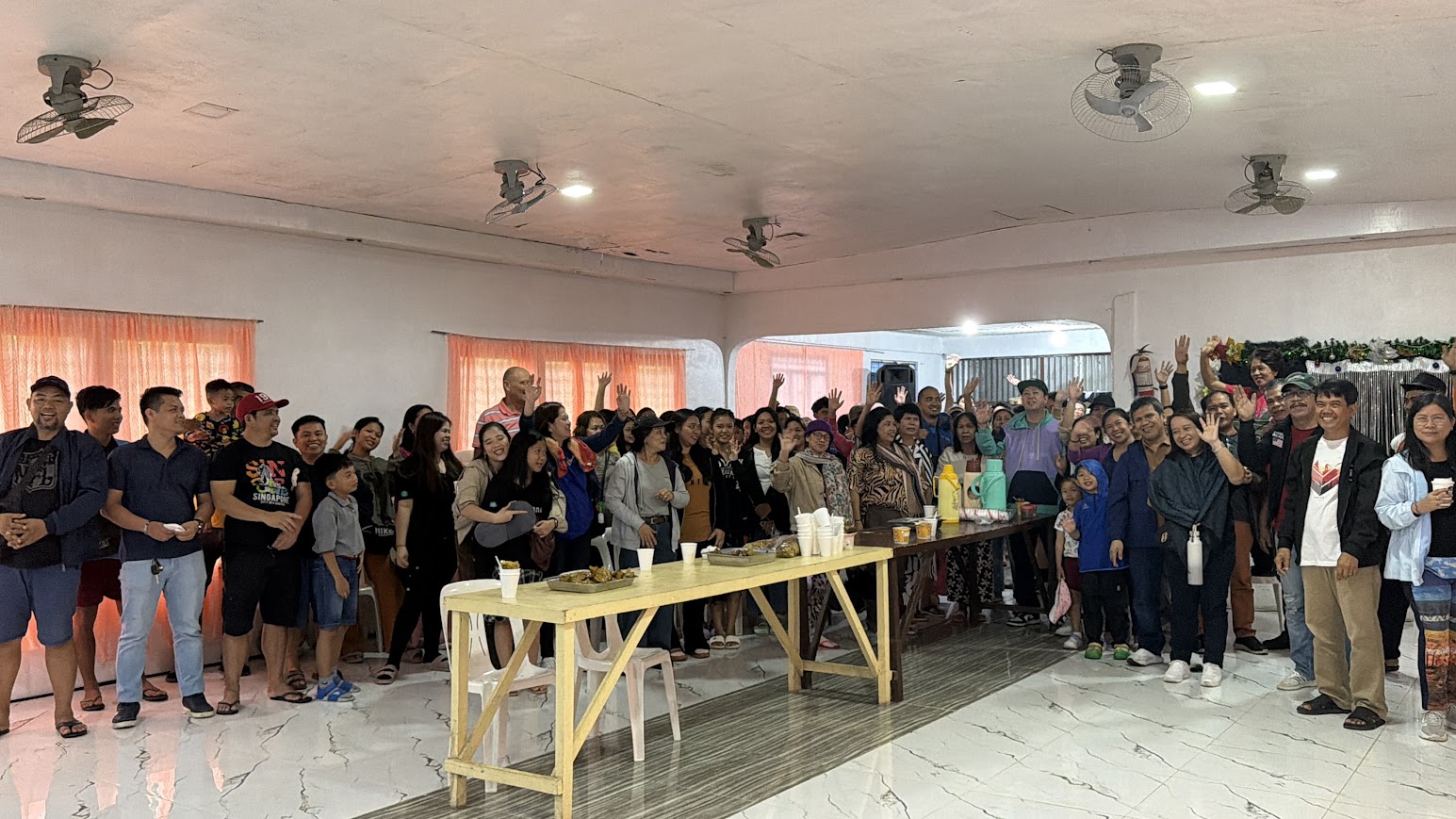 Morning fellowship — the crowd waves from behind a long table of food.